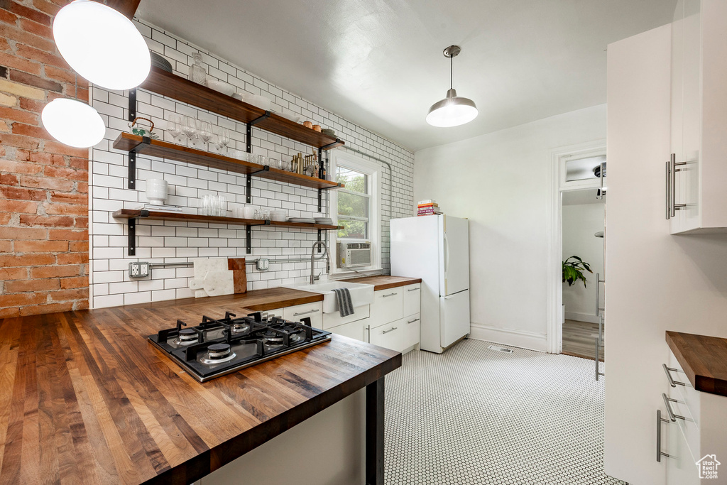 Kitchen with wooden counters, stainless steel gas cooktop, freestanding refrigerator, white cabinets, and brick wall