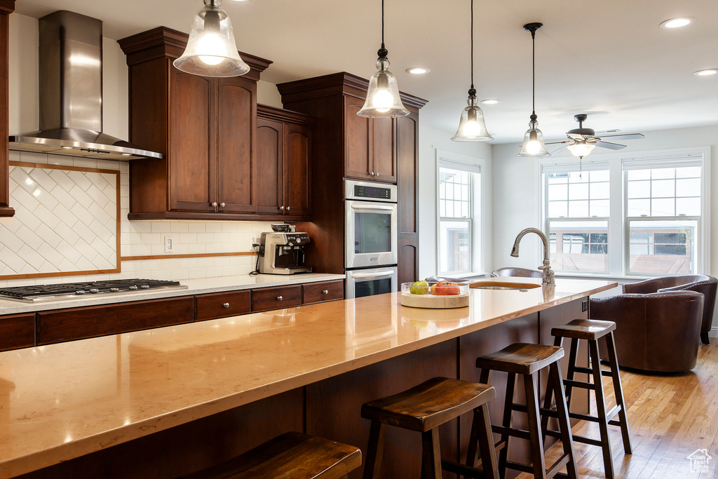 Kitchen with wall chimney range hood, stainless steel appliances, light stone countertops, backsplash, and a ceiling fan