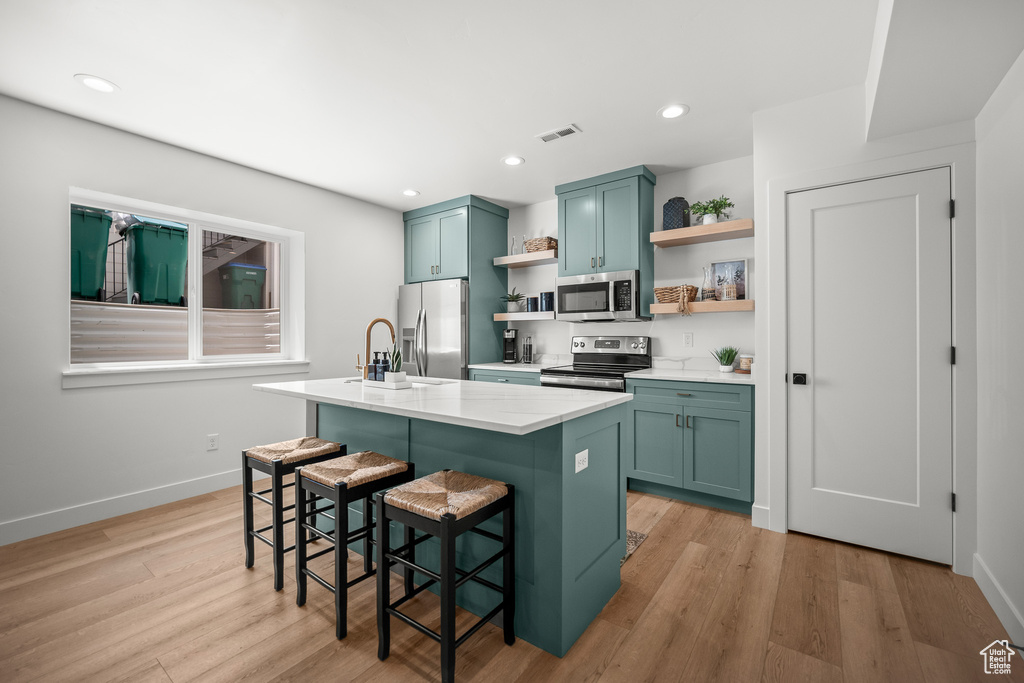 Kitchen featuring open shelves, stainless steel appliances, a kitchen island with sink, light wood finished floors, and a breakfast bar