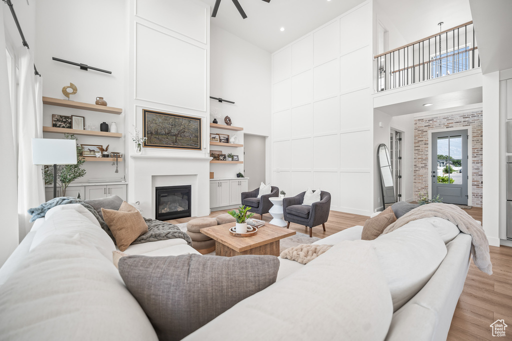 Living area with a high ceiling, a glass covered fireplace, light wood finished floors, a ceiling fan, and built in features