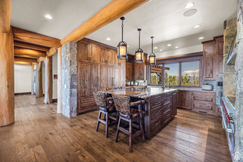 Kitchen with dark wood-type flooring, recessed lighting, an island with sink, beam ceiling, and a breakfast bar