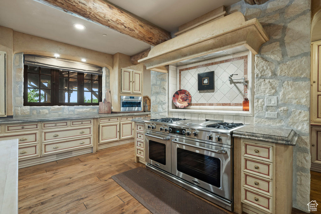 Kitchen with range with two ovens, decorative backsplash, light wood-style floors, cream cabinetry, and beamed ceiling