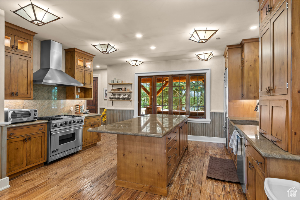 Kitchen with designer stove, wall chimney exhaust hood, a center island, brown cabinets, and a wainscoted wall