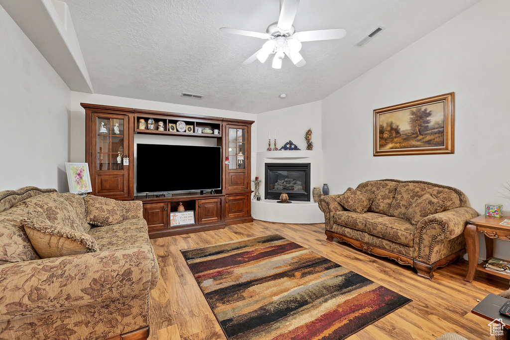 Living room with light wood-type flooring, ceiling fan, a glass covered fireplace, and a textured ceiling