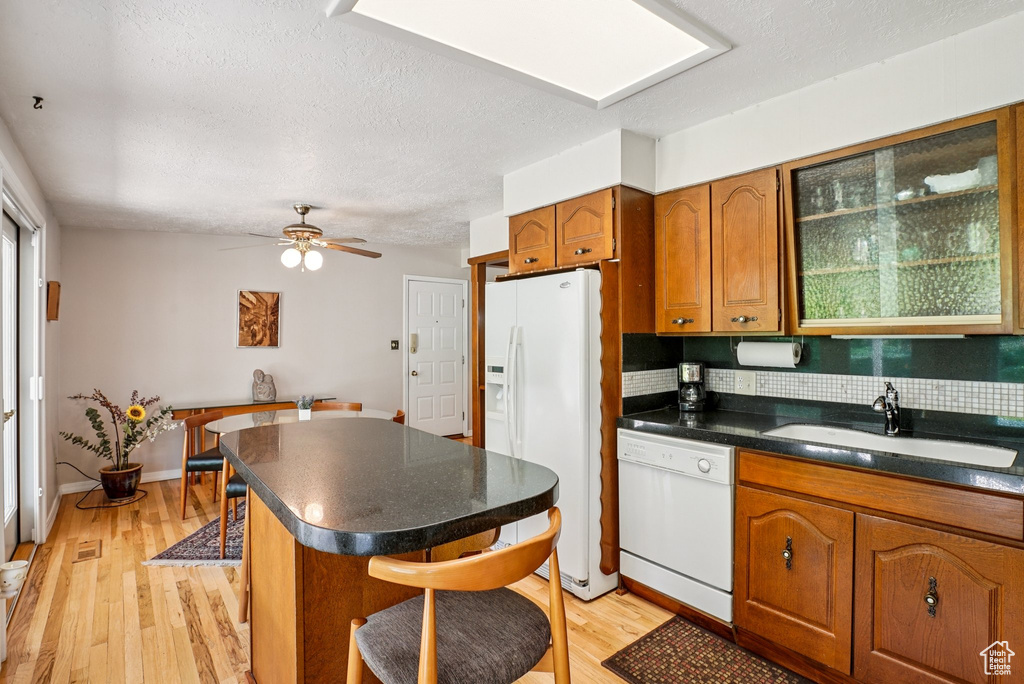 Kitchen featuring white appliances, dark countertops, brown cabinetry, a kitchen bar, and light wood-type flooring