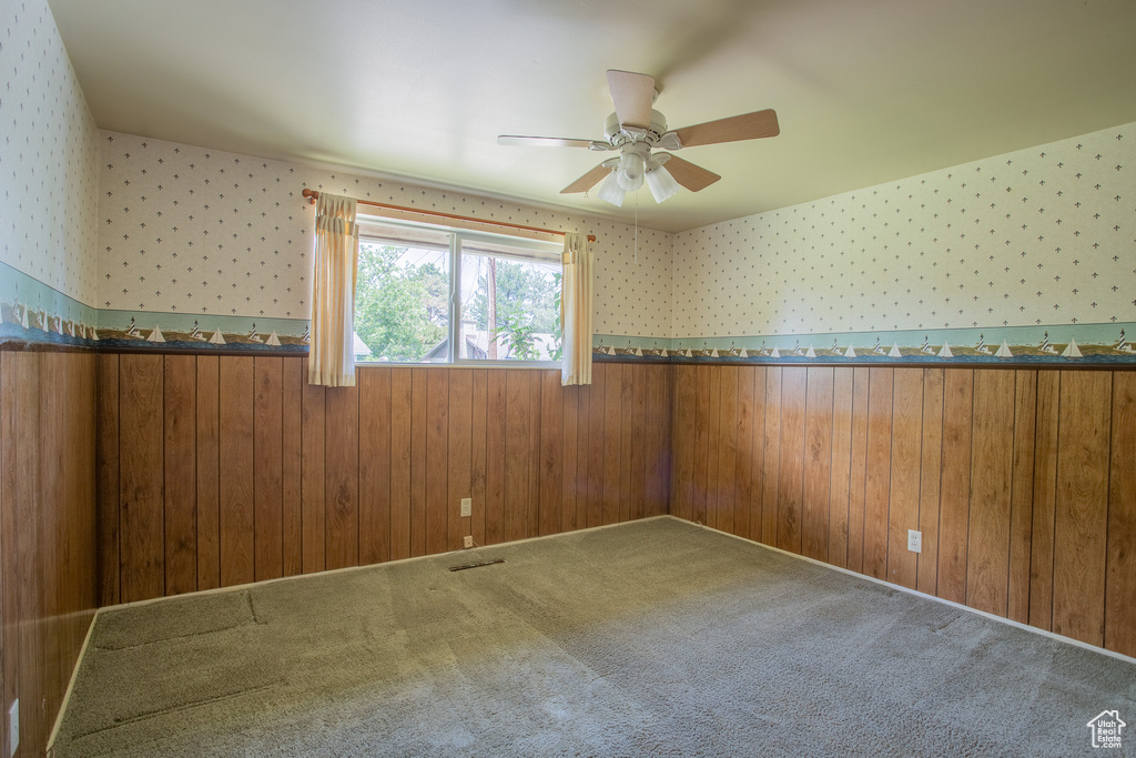 Carpeted empty room featuring a wainscoted wall, wallpapered walls, a ceiling fan, and wooden walls