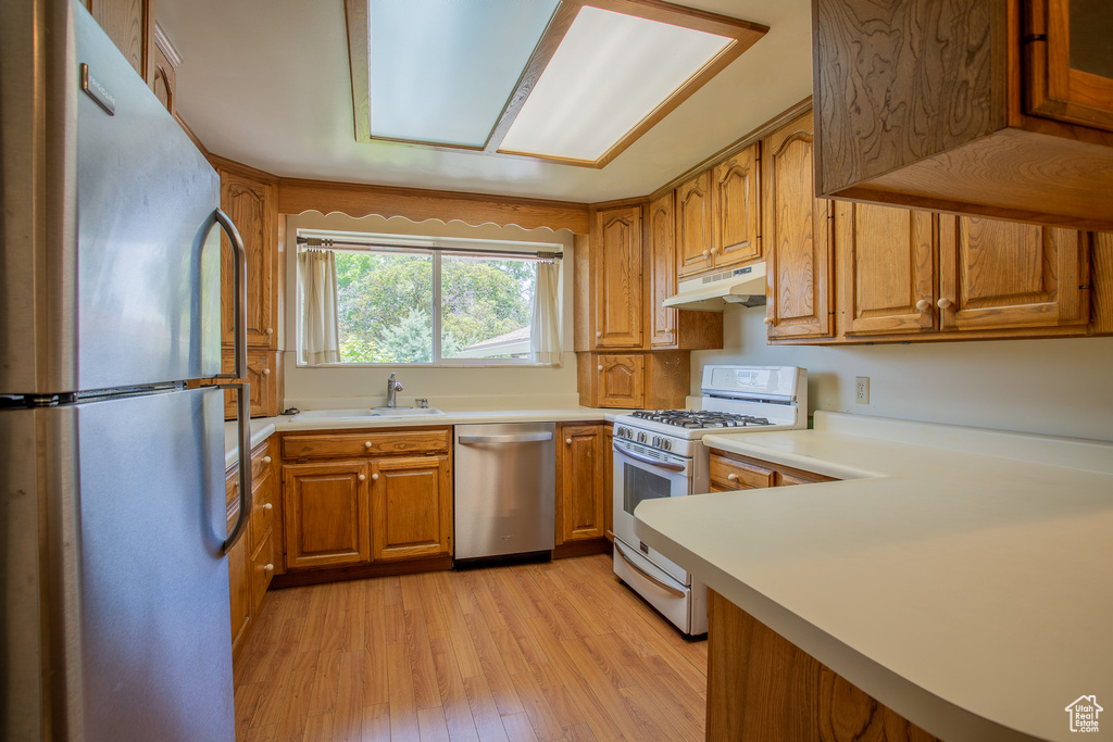 Kitchen with stainless steel appliances, under cabinet range hood, light wood-type flooring, brown cabinets, and light countertops