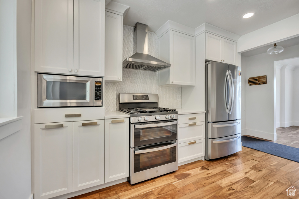 Kitchen featuring stainless steel appliances, wall chimney exhaust hood, tasteful backsplash, light wood-style floors, and light countertops
