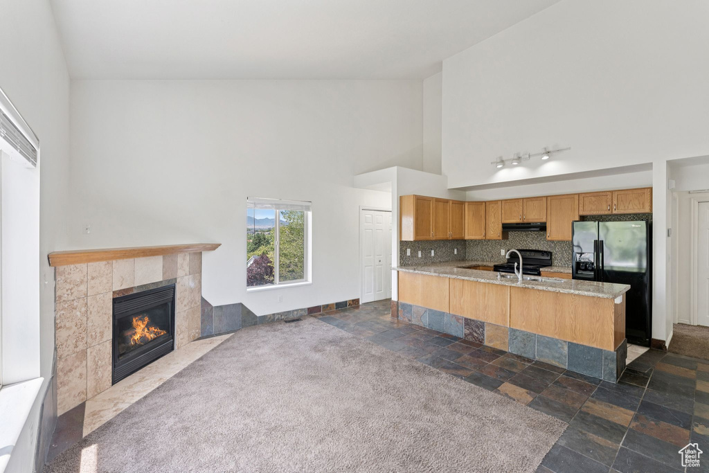 Kitchen with black fridge with ice dispenser, a tile fireplace, high vaulted ceiling, open floor plan, and under cabinet range hood