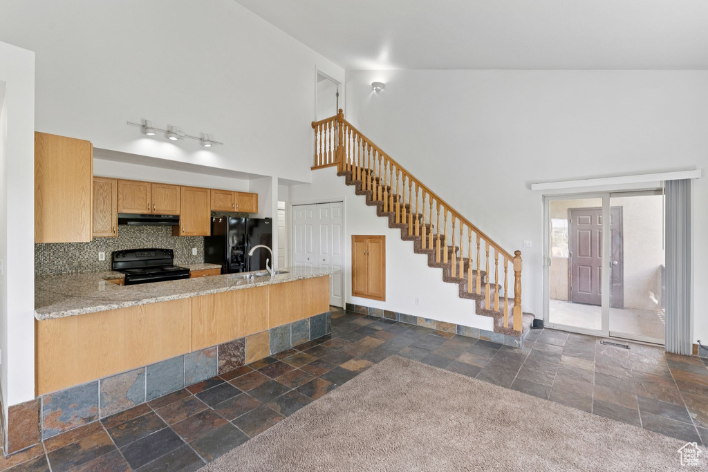Kitchen with black appliances, stone tile floors, a towering ceiling, a peninsula, and under cabinet range hood