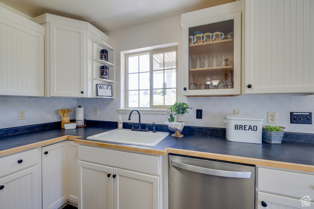 Kitchen featuring stainless steel dishwasher, dark countertops, backsplash, glass insert cabinets, and white cabinetry