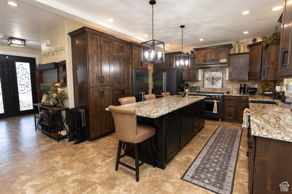 Kitchen with black appliances, decorative backsplash, a center island with sink, light stone countertops, and dark brown cabinetry