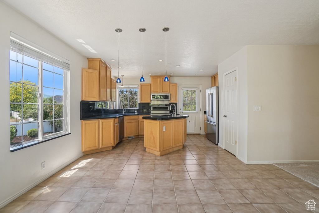 Kitchen featuring stainless steel appliances, an island with sink, dark countertops, light tile patterned floors, and hanging light fixtures
