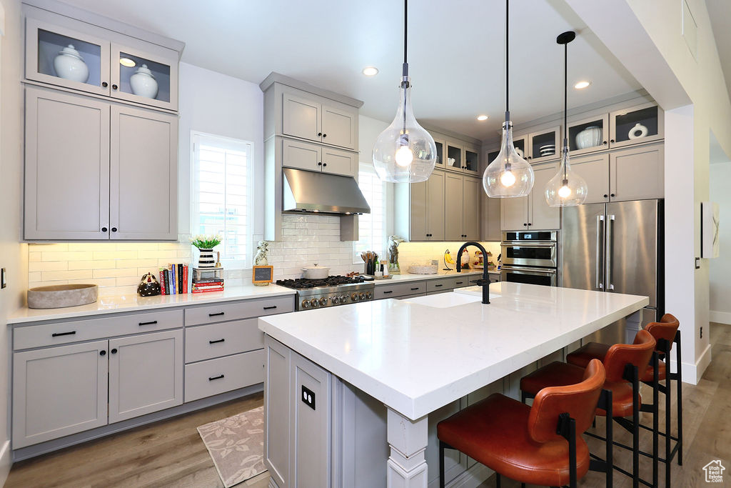 Kitchen with under cabinet range hood, light wood finished floors, a kitchen island with sink, decorative backsplash, and a breakfast bar