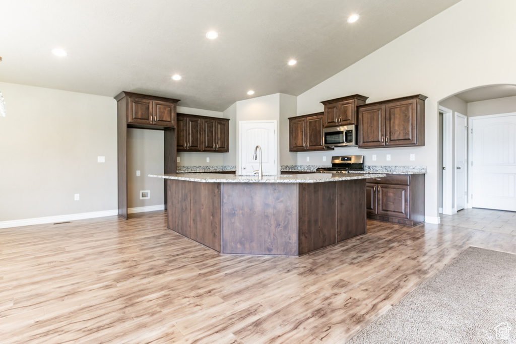Kitchen with stainless steel appliances, a large island with sink, dark brown cabinets, light wood-style floors, and arched walkways