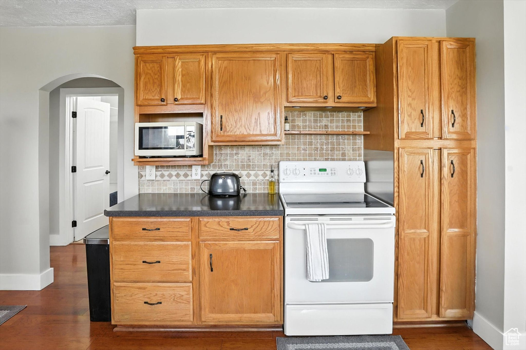 Kitchen featuring white appliances, arched walkways, dark wood-style floors, backsplash, and dark countertops