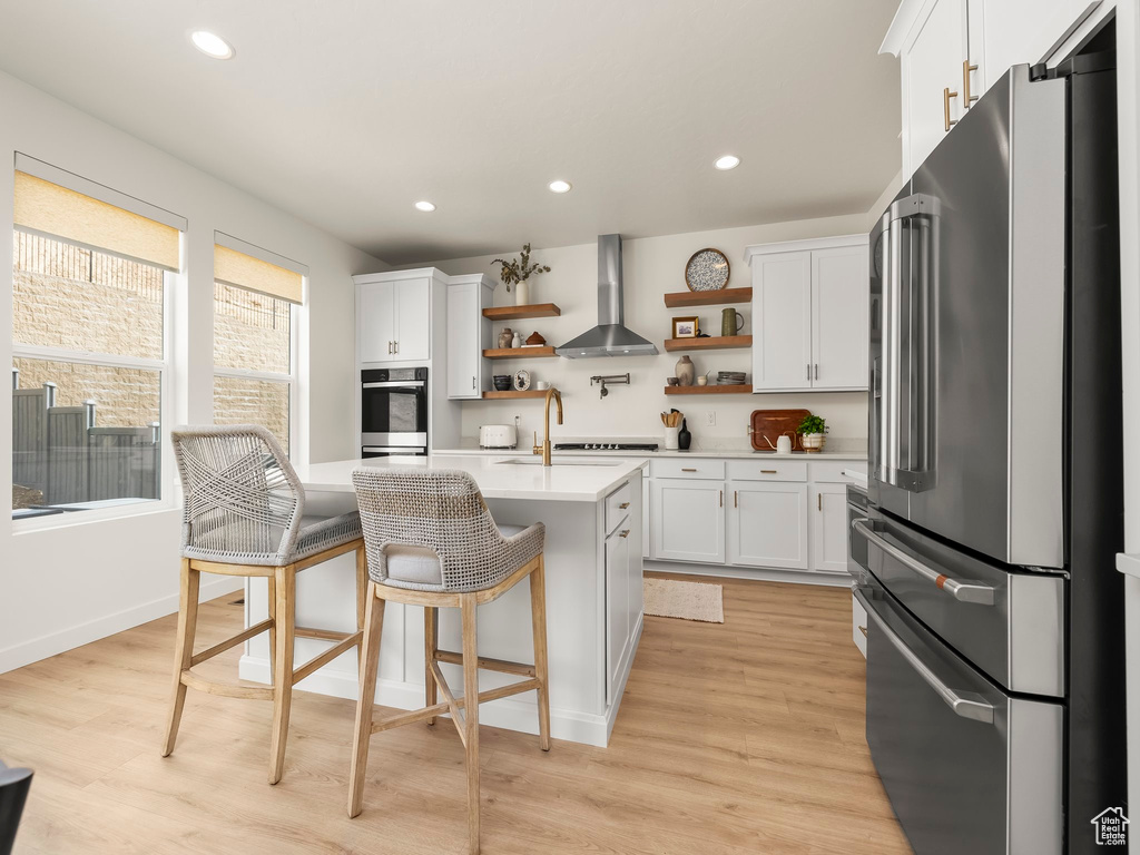 Kitchen with high end refrigerator, wall chimney range hood, open shelves, light countertops, and light wood-type flooring