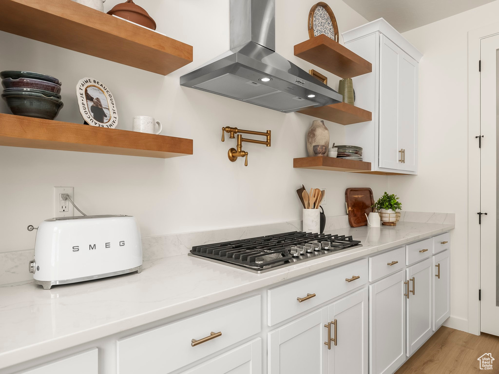 Kitchen with open shelves, wall chimney range hood, stainless steel gas stovetop, white cabinets, and light countertops