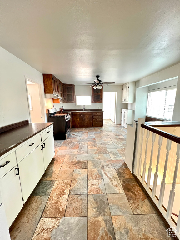 Kitchen featuring gas stove, stone tile flooring, ceiling fan, dark countertops, and under cabinet range hood