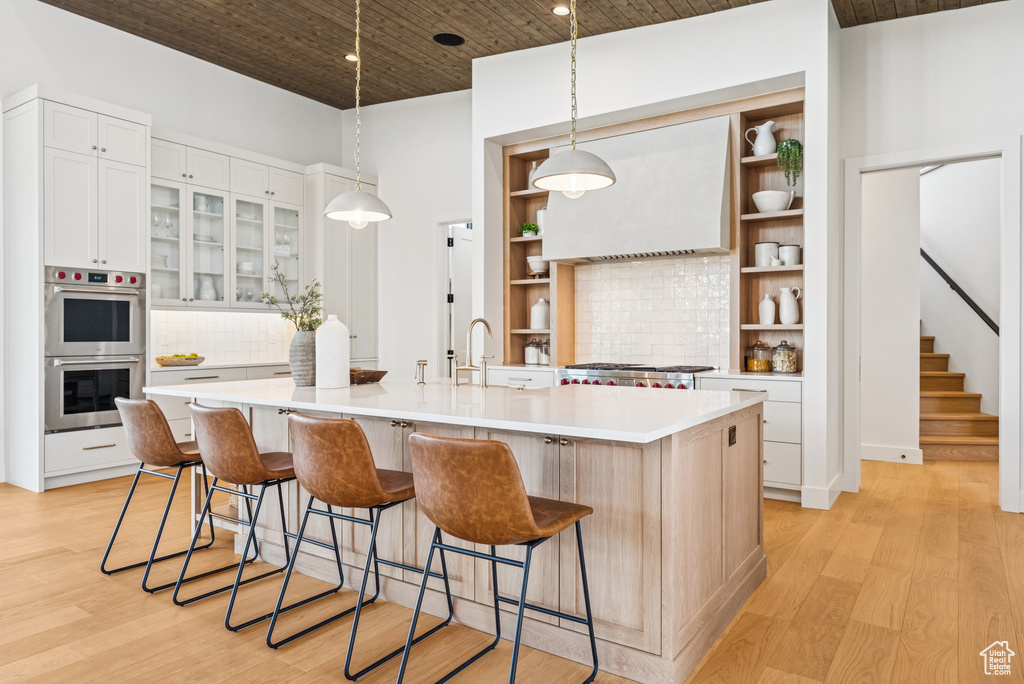Kitchen featuring double oven, ventilation hood, open shelves, light countertops, and light wood finished floors
