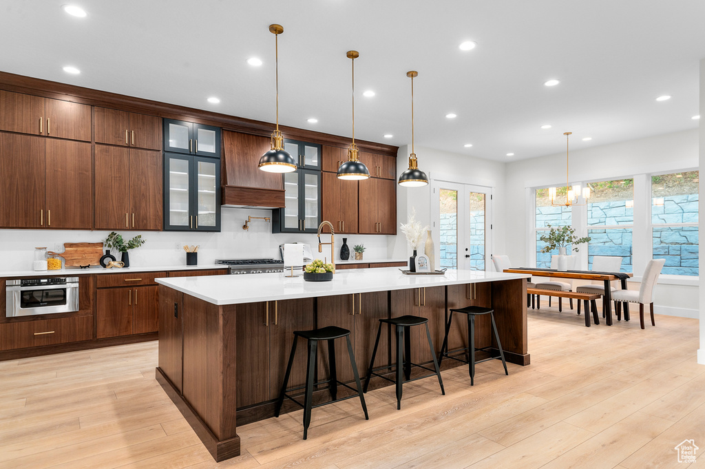 Kitchen featuring light wood finished floors, a large island with sink, light countertops, recessed lighting, and glass insert cabinets