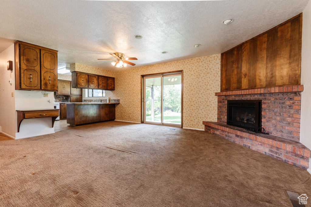 Unfurnished living room featuring a ceiling fan, a brick fireplace, light colored carpet, wallpapered walls, and a textured ceiling