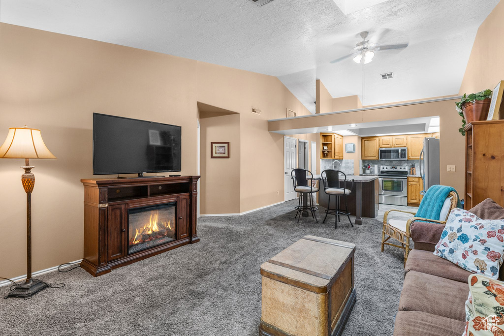 Living area featuring vaulted ceiling, light carpet, a ceiling fan, a glass covered fireplace, and a textured ceiling