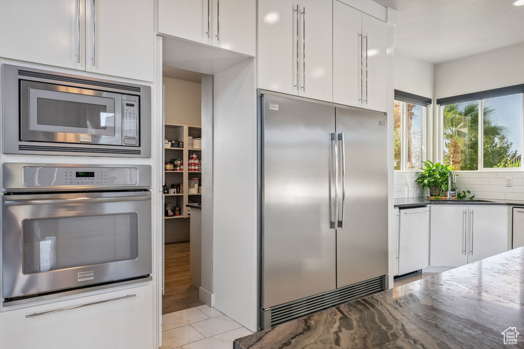 Kitchen featuring built in appliances, white cabinets, tasteful backsplash, dark stone countertops, and modern cabinets