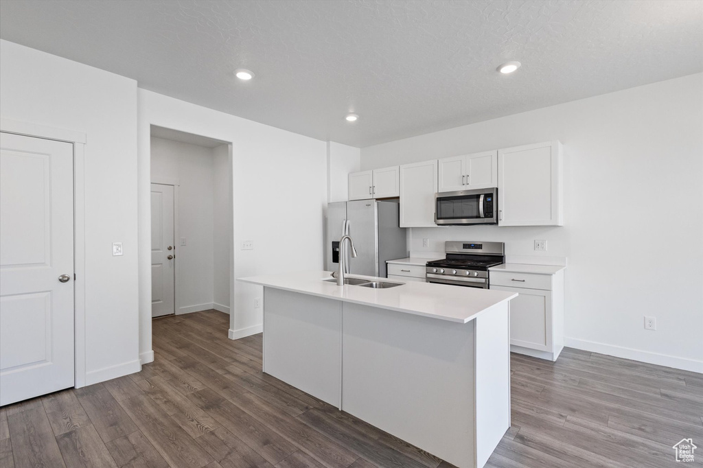 Kitchen featuring stainless steel appliances, white cabinets, wood finished floors, a kitchen island with sink, and light countertops