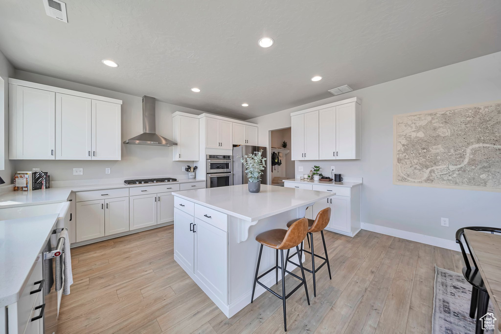 Kitchen featuring wall chimney exhaust hood, appliances with stainless steel finishes, a kitchen bar, a kitchen island, and white cabinetry