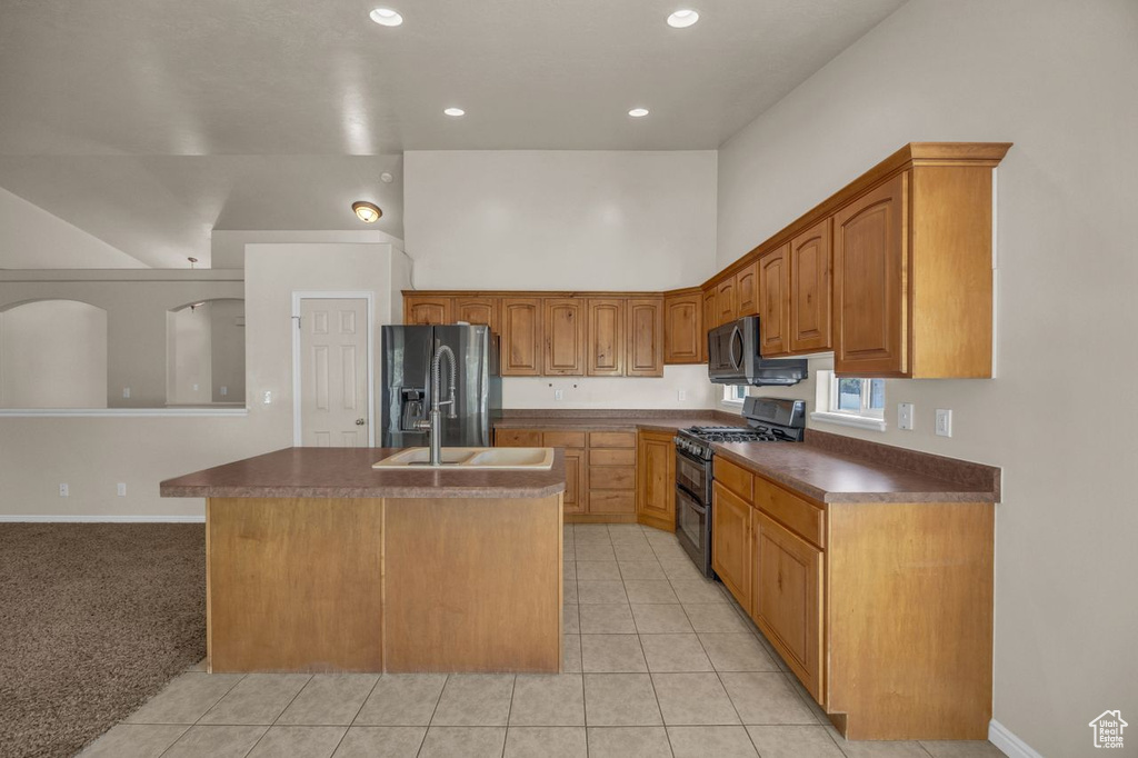 Kitchen with double oven range, refrigerator with ice dispenser, a kitchen island with sink, light tile patterned floors, and dark countertops
