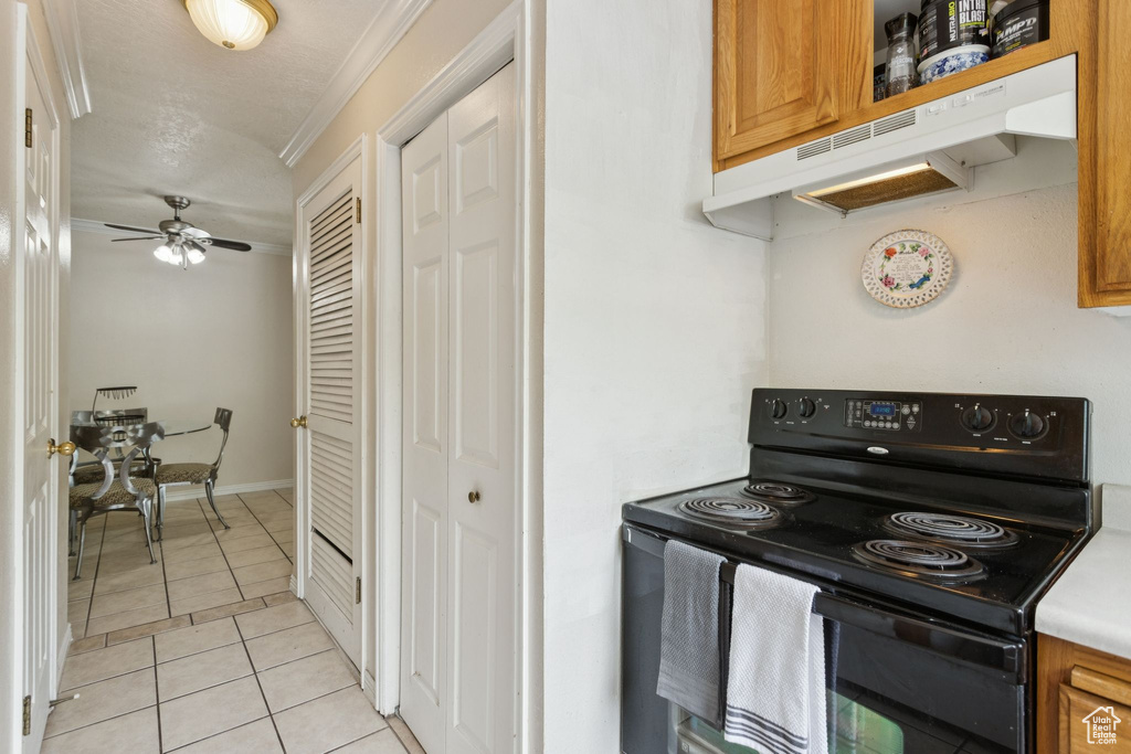 Kitchen featuring black / electric stove, under cabinet range hood, brown cabinetry, light tile patterned flooring, and ceiling fan