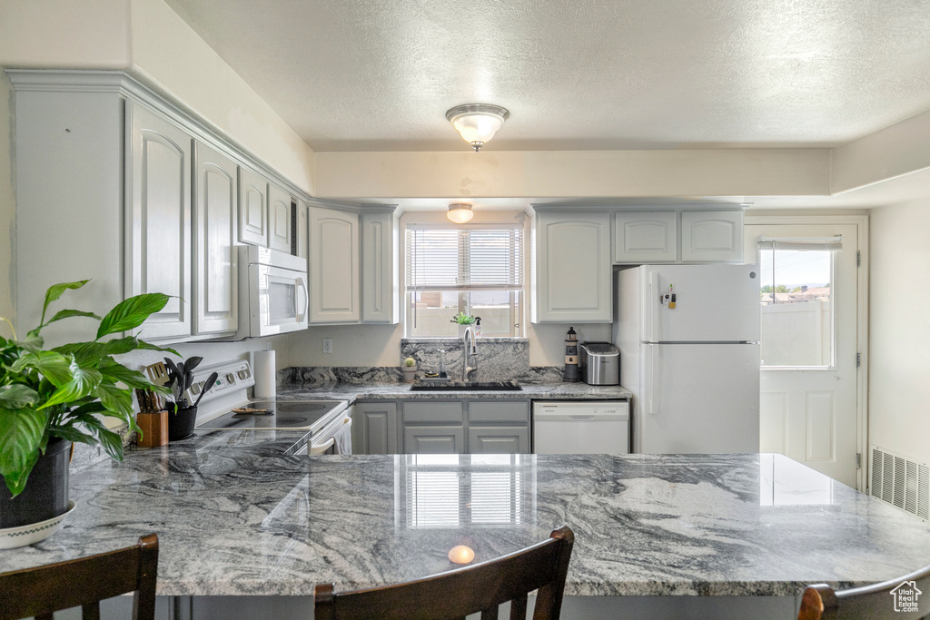 Kitchen with white appliances, a peninsula, a breakfast bar, a textured ceiling, and light stone counters