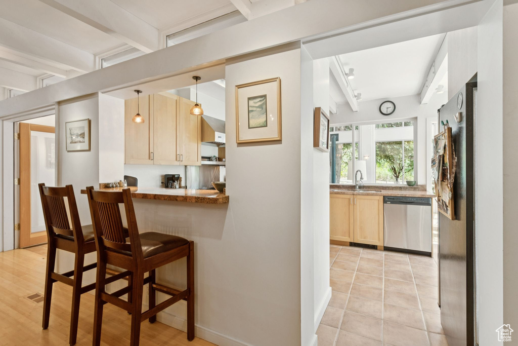 Kitchen featuring appliances with stainless steel finishes, light brown cabinetry, beamed ceiling, a kitchen bar, and pendant lighting