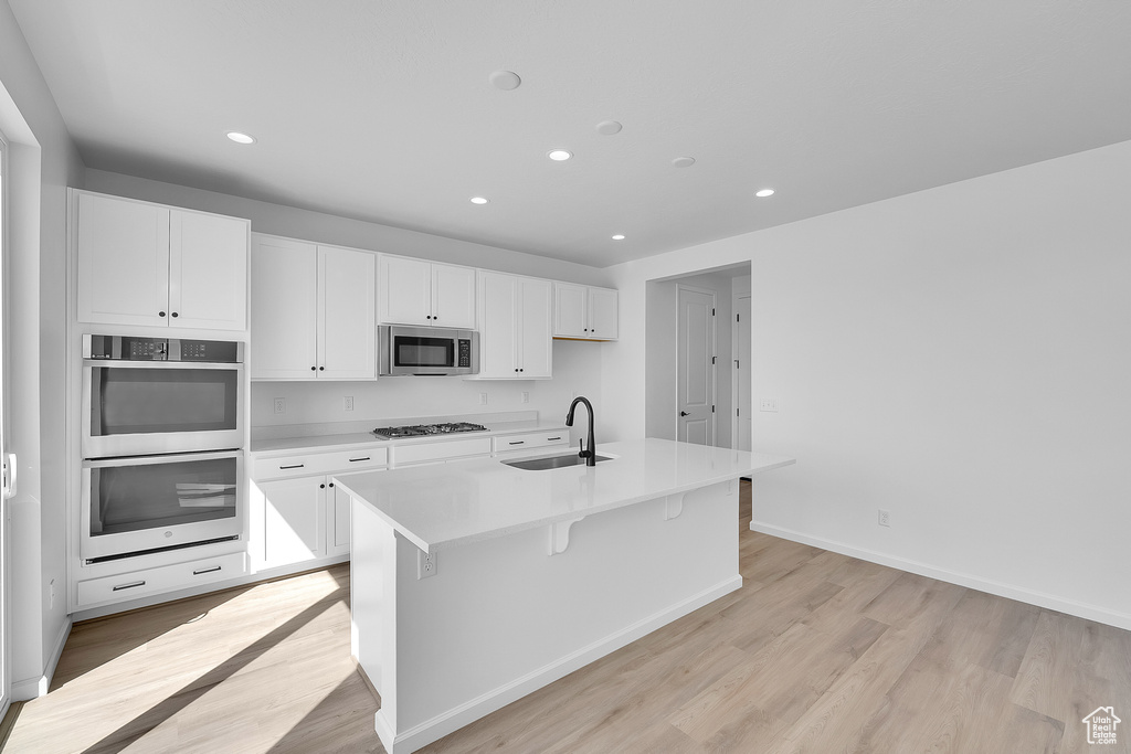 Kitchen with stainless steel appliances, light wood-style floors, white cabinetry, recessed lighting, and a center island with sink