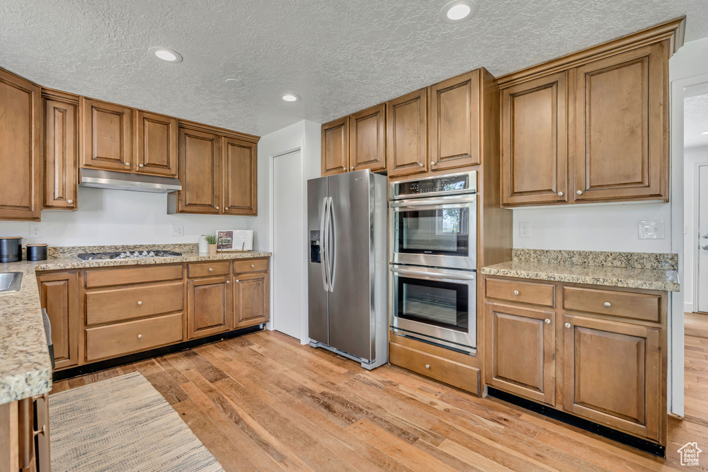 Kitchen featuring appliances with stainless steel finishes, light wood finished floors, brown cabinets, under cabinet range hood, and a textured ceiling