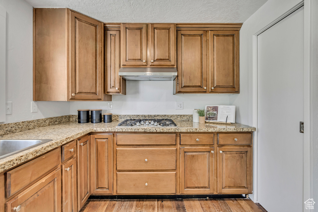 Kitchen featuring under cabinet range hood, stainless steel gas stovetop, light wood finished floors, brown cabinets, and a textured ceiling