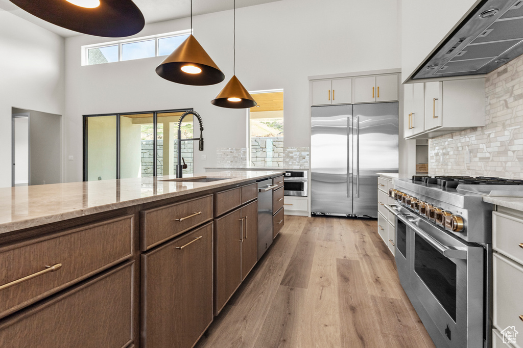 Kitchen with premium appliances, range hood, a high ceiling, light wood-style flooring, and tasteful backsplash