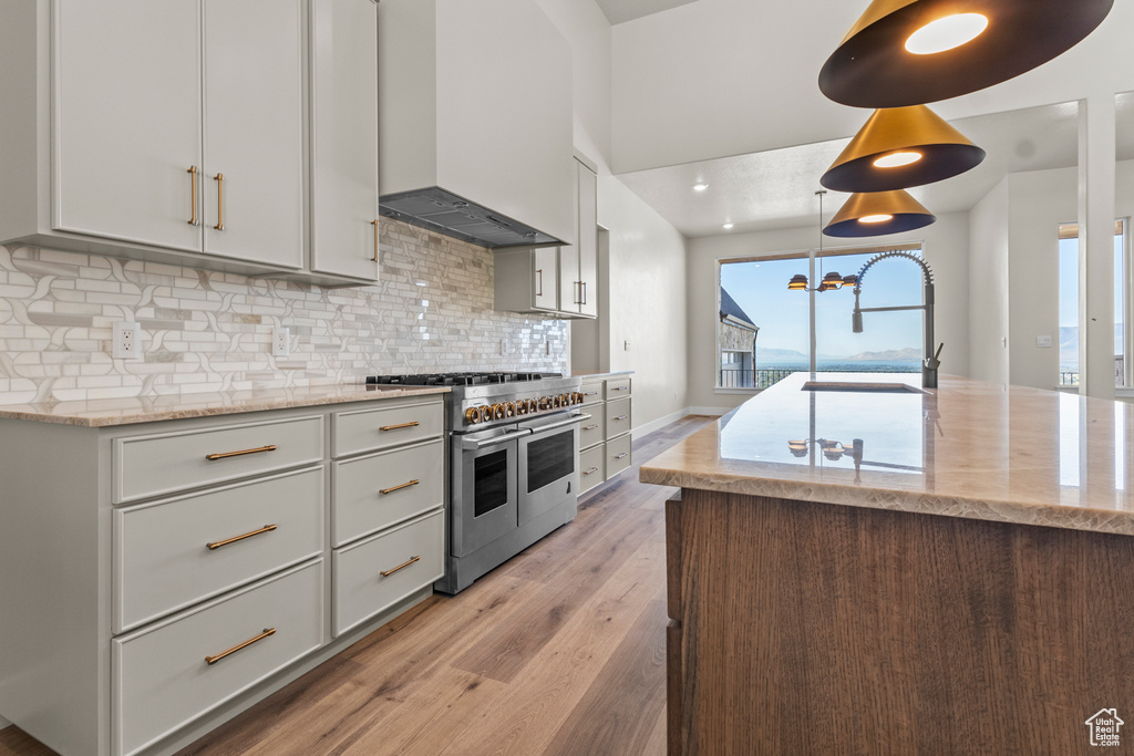 Kitchen featuring range with two ovens, custom exhaust hood, light wood finished floors, backsplash, and light stone countertops
