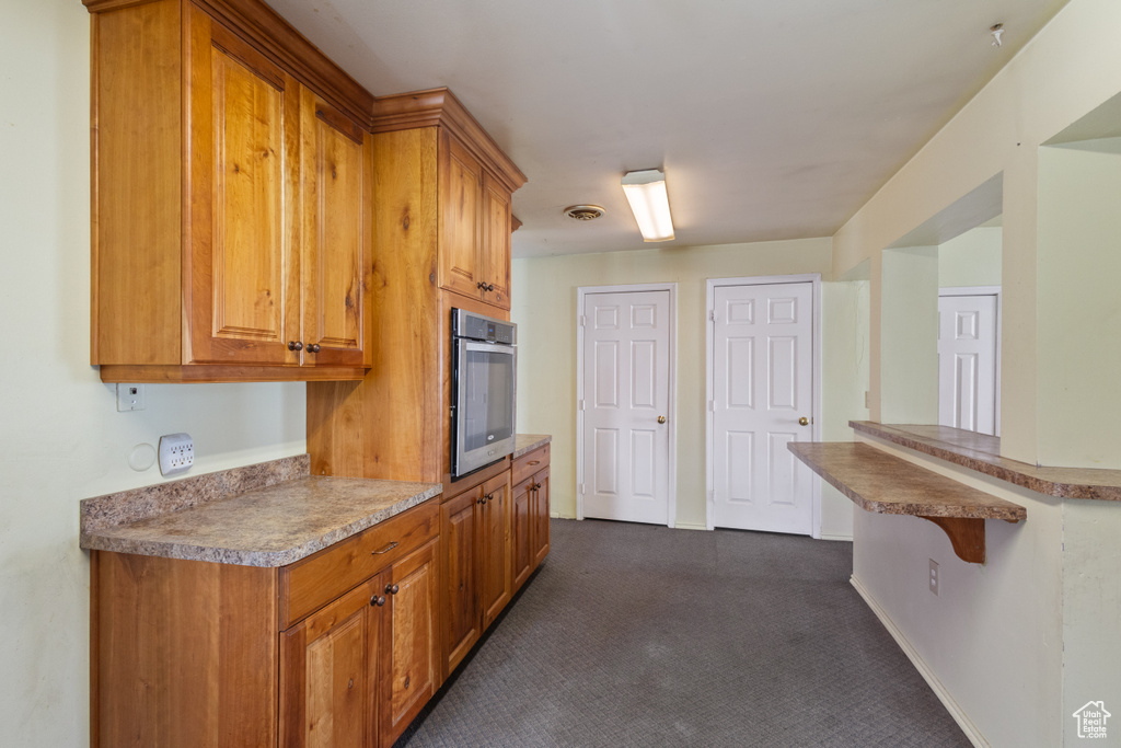Kitchen with stainless steel oven, dark colored carpet, and brown cabinets