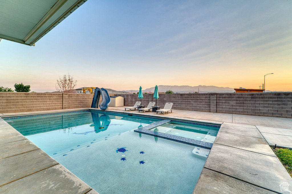 Pool at dusk featuring a water slide, a patio area, a fenced backyard, and a pool with connected hot tub