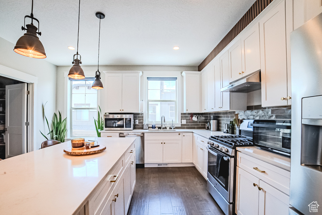Kitchen with under cabinet range hood, stainless steel appliances, light countertops, backsplash, and dark wood-style floors