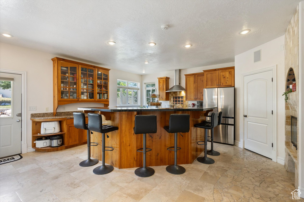 Kitchen with freestanding refrigerator, brown cabinetry, wall chimney range hood, glass insert cabinets, and a breakfast bar area