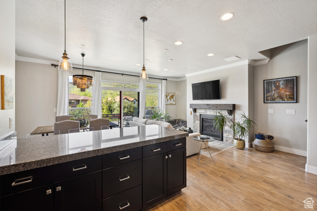 Kitchen featuring dark cabinets, crown molding, a textured ceiling, a stone fireplace, and light wood-style floors