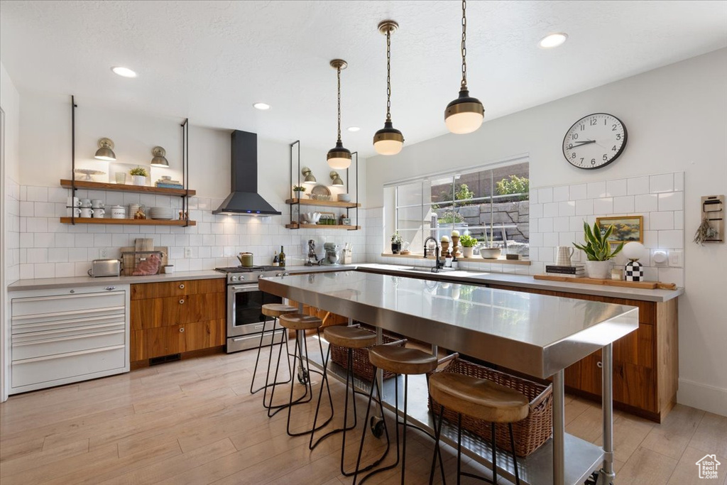 Kitchen featuring wall chimney exhaust hood, stainless steel range with gas cooktop, brown cabinetry, open shelves, and light wood-style floors