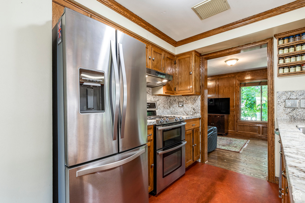 Kitchen featuring stainless steel appliances, under cabinet range hood, brown cabinetry, decorative backsplash, and ornamental molding