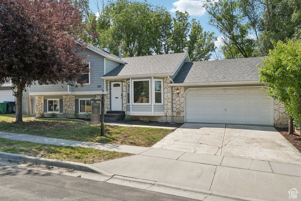 Tri-level home featuring stone siding, a garage, driveway, roof with shingles, and a front yard