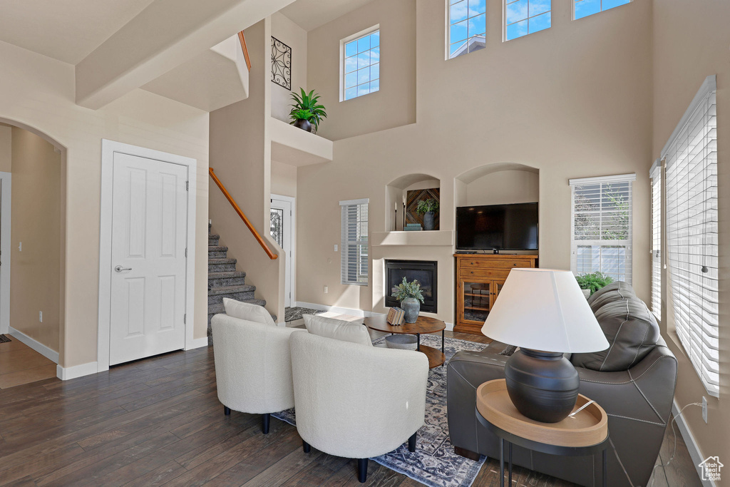 Living area featuring dark wood finished floors, a glass covered fireplace, healthy amount of natural light, stairway, and a towering ceiling