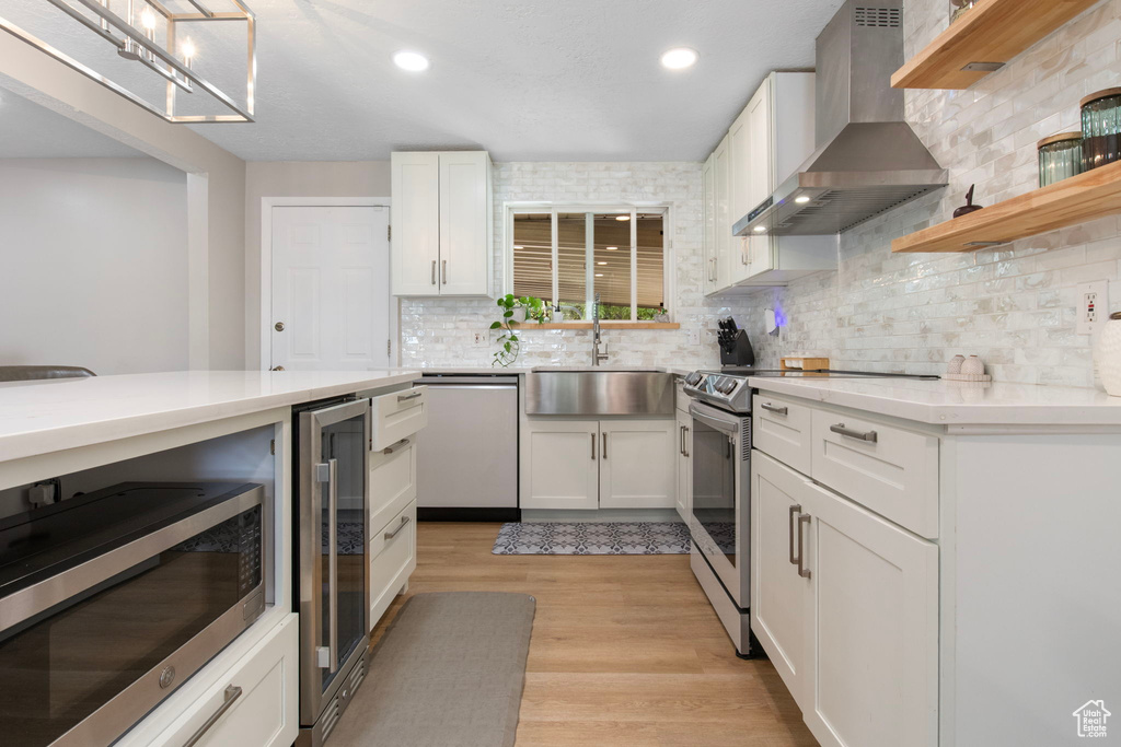 Kitchen featuring wall chimney range hood, appliances with stainless steel finishes, beverage cooler, light countertops, and white cabinetry