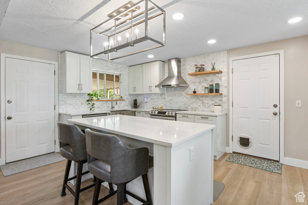 Kitchen with wall chimney exhaust hood, light wood-style floors, stainless steel range with electric cooktop, light countertops, and a textured ceiling